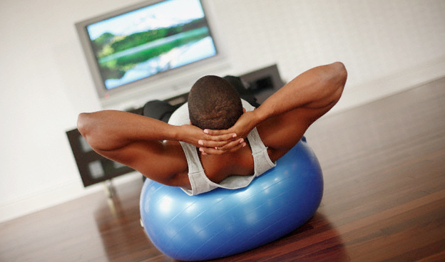 A man watching tv while doing crunches on a blue fitness ball