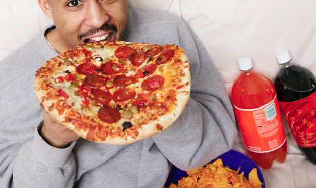 A man biting into a whole pizza while sitting on his sofa
