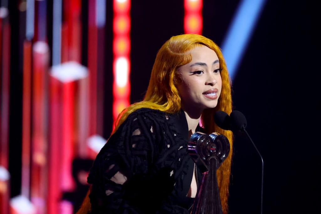 Ice Spice with long orange hair, wearing a black textured top, holding a silver award trophy, and speaking into microphones on a stage with red and blue lighting.
