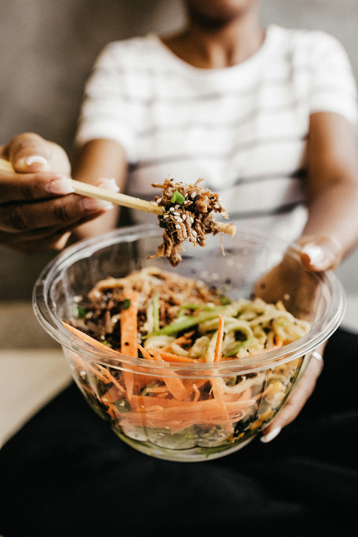 Close-up of hands holding chopsticks lifting shredded meat with sesame seeds from a clear bowl containing a colorful meal with carrots and green vegetables.