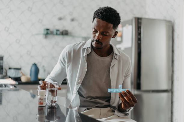 A Black man in a kitchen holds a blue pill organizer, looking down at it. A glass of water and a pill bottle are on the counter beside him.