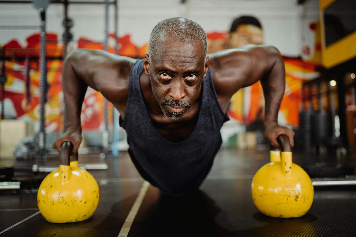 A muscular older Black man with a graying mustache and goatee, wearing a dark tank top, performs a push-up using two yellow kettlebells as handles. He looks directly at the camera with an intense expression in a gym setting.