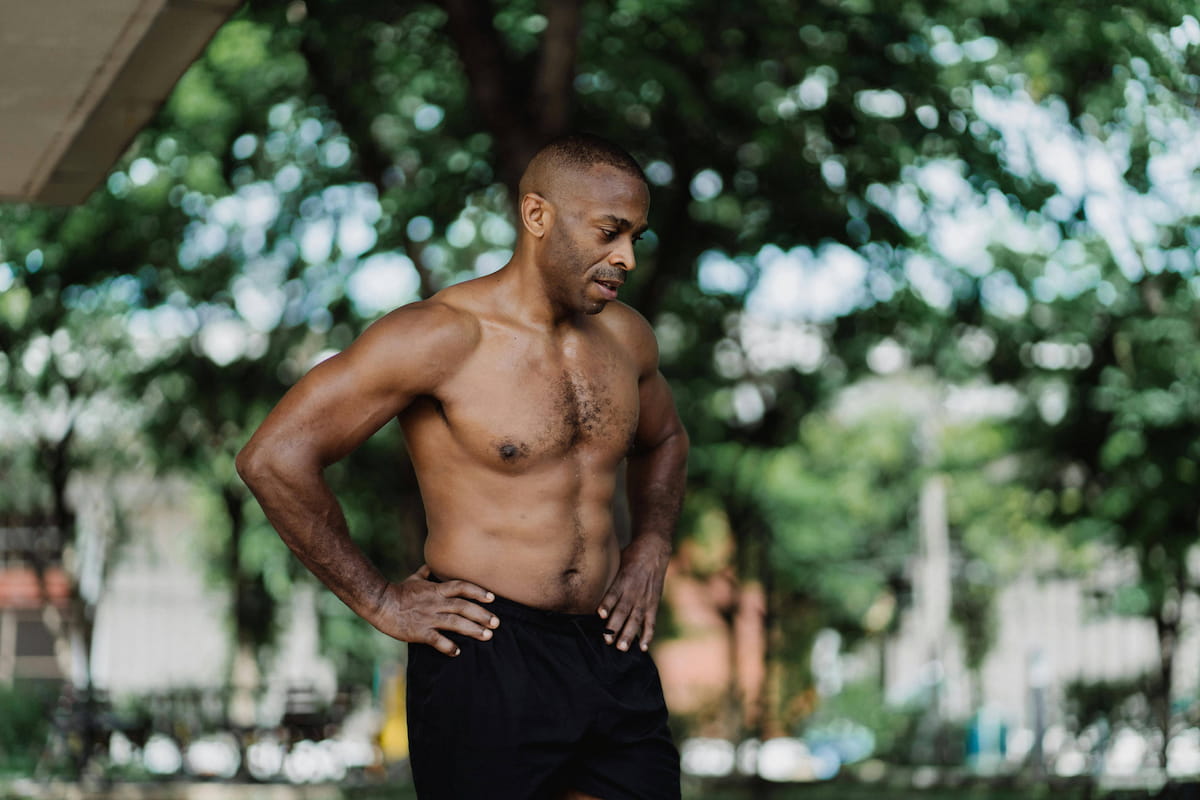 A shirtless Black man with a muscular build stands with hands on hips, looking down, against a blurred green outdoor background.