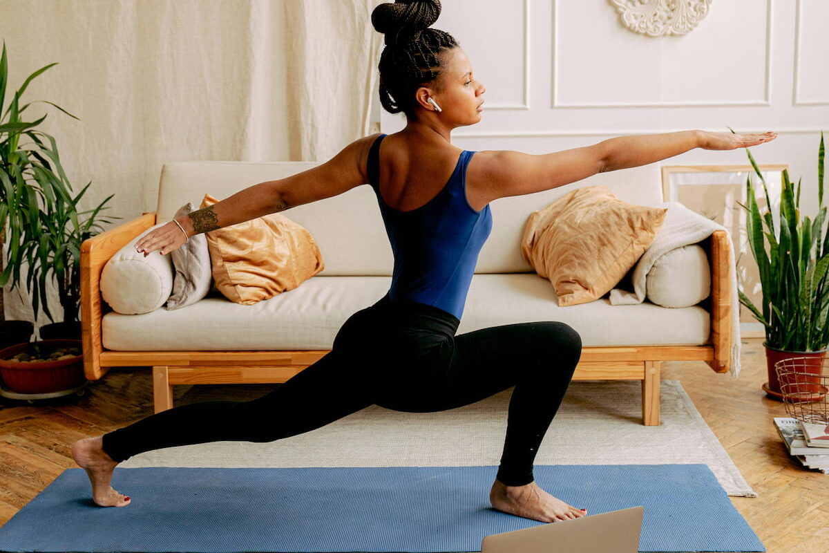 A woman in a blue leotard and black leggings practices yoga on a blue mat at home, following a class on a laptop. She is in a Warrior II pose, with arms extended and looking forward. A sofa, plants, and decorative wall art are in the background.