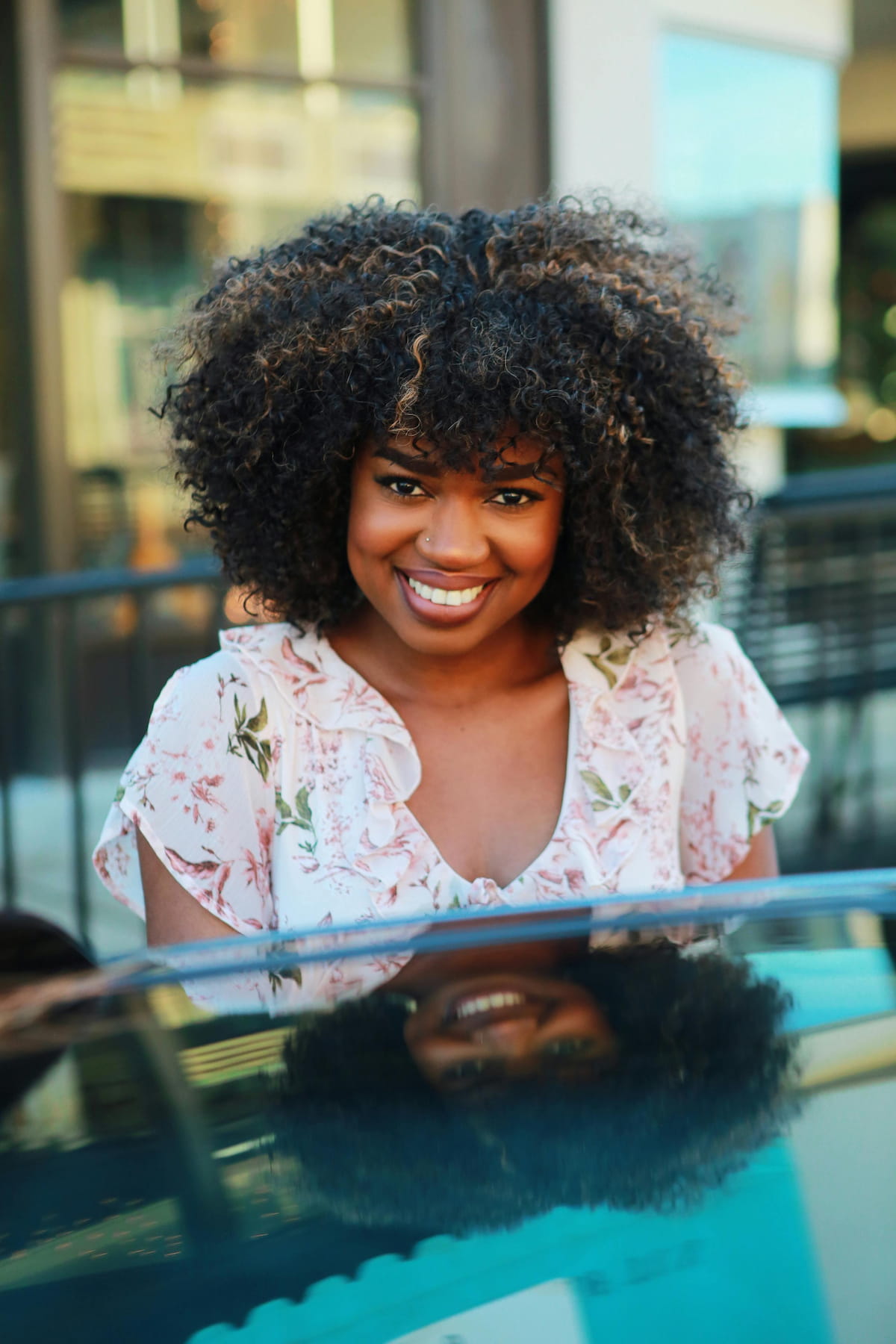 A smiling Black woman with voluminous curly hair and a floral top, reflected in a dark surface.
