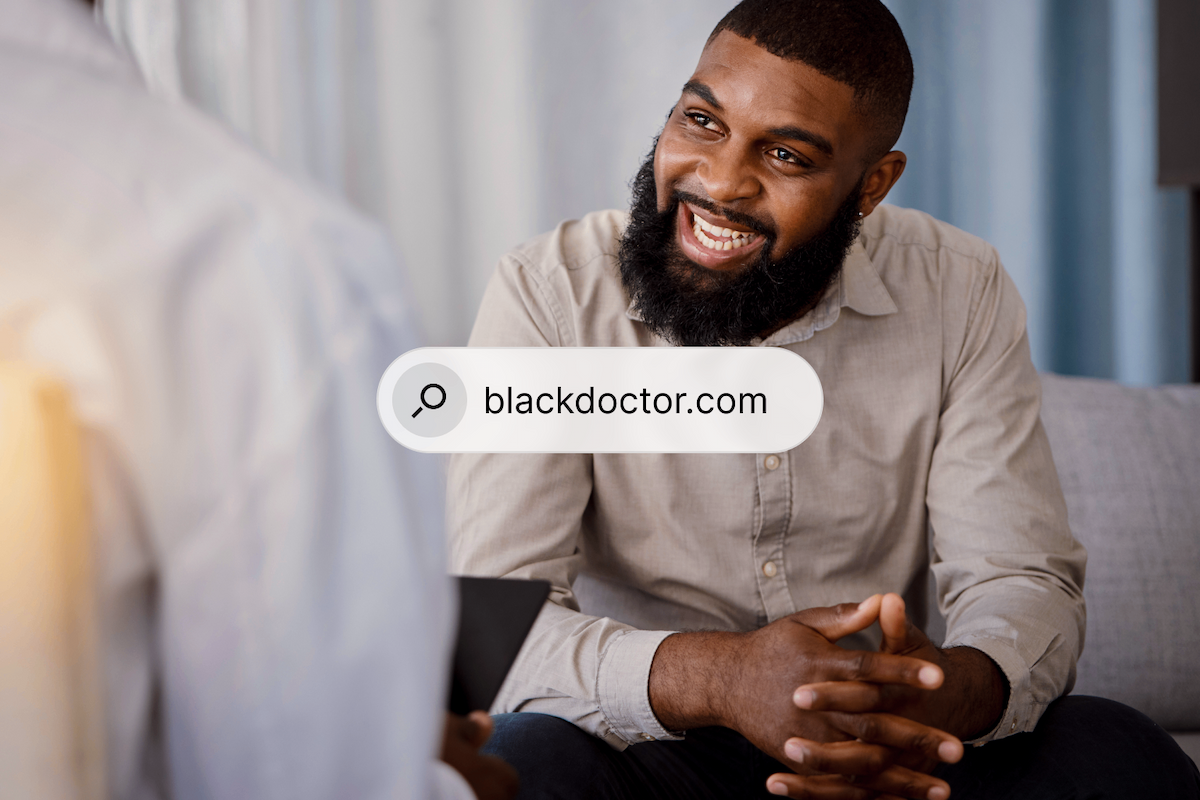 A Black man with a beard smiles broadly while looking at a person in a white coat, with a search bar overlay showing 'blackdoctor.com'.